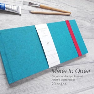 May include: A teal sketchbook with a red elastic band closure. The cover has a textured appearance and a white label with the text "Printedin Press & Bindery". The image also shows a tube of blue paint and two paintbrushes.