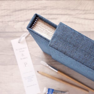 May include: A blue fabric-covered journal with a spiral binding, accompanied by a white bookmark featuring the text "Printblock Press & Bindery & P", a wooden pencil, a paintbrush with a wooden handle, and a small tube of blue paint.