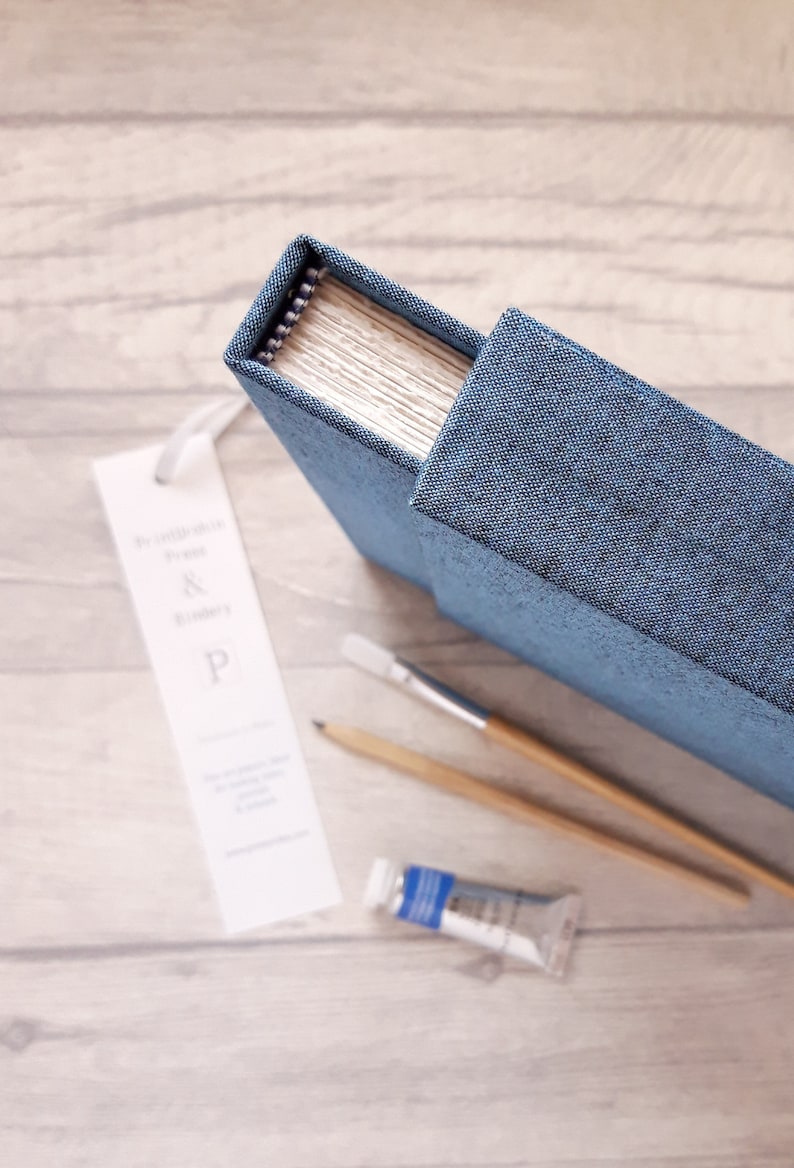 May include: A blue fabric-covered journal with a spiral binding, accompanied by a white bookmark featuring the text "Printblock Press & Bindery & P", a wooden pencil, a paintbrush with a wooden handle, and a small tube of blue paint.