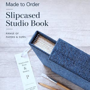 May include: A blue fabric-covered slipcased studio book with visible paper edges, accompanied by a white bookmark labeled PrintUrchin Press & Bindery, a wooden pencil, and a paintbrush. Text reads Made to Order, Slipcased Studio Book.