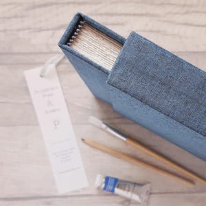 May include: A blue fabric-covered sketchbook with a spiral binding, accompanied by a white bookmark featuring the text "& P", a wooden pencil, a paintbrush with a wooden handle, and a small tube of blue paint, all arranged on a light surface.