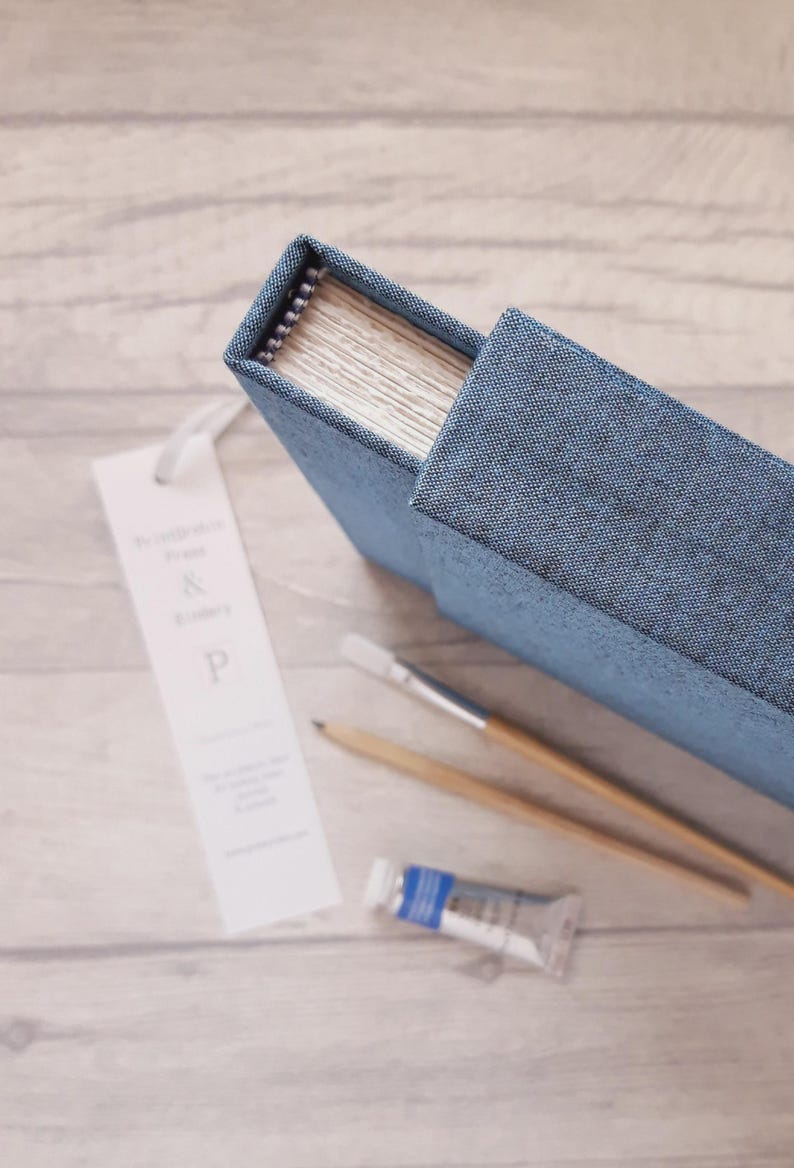 May include: A blue fabric-covered sketchbook with a spiral binding, accompanied by a white bookmark featuring the text "& P", a wooden pencil, a paintbrush with a wooden handle, and a small tube of blue paint, all arranged on a light surface.