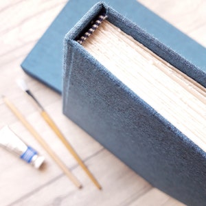 May include: A hardbound book with a textured blue fabric cover and cream-colored pages. The spine features a small black and white patterned fabric detail at the top. The book is positioned near two thin wooden paintbrushes and a small paint tube.