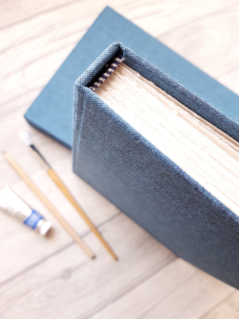 May include: A hardbound book with a textured blue fabric cover and cream-colored pages. The spine features a small black and white patterned fabric detail at the top. The book is positioned near two thin wooden paintbrushes and a small paint tube.