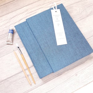 May include: A blue fabric-covered journal with a white ribbon bookmark featuring the text Printurchin Press & Bindery P. Beside the journal are a small tube of blue paint, a wooden paintbrush, and a wooden pencil, all arranged on a light wood surface.