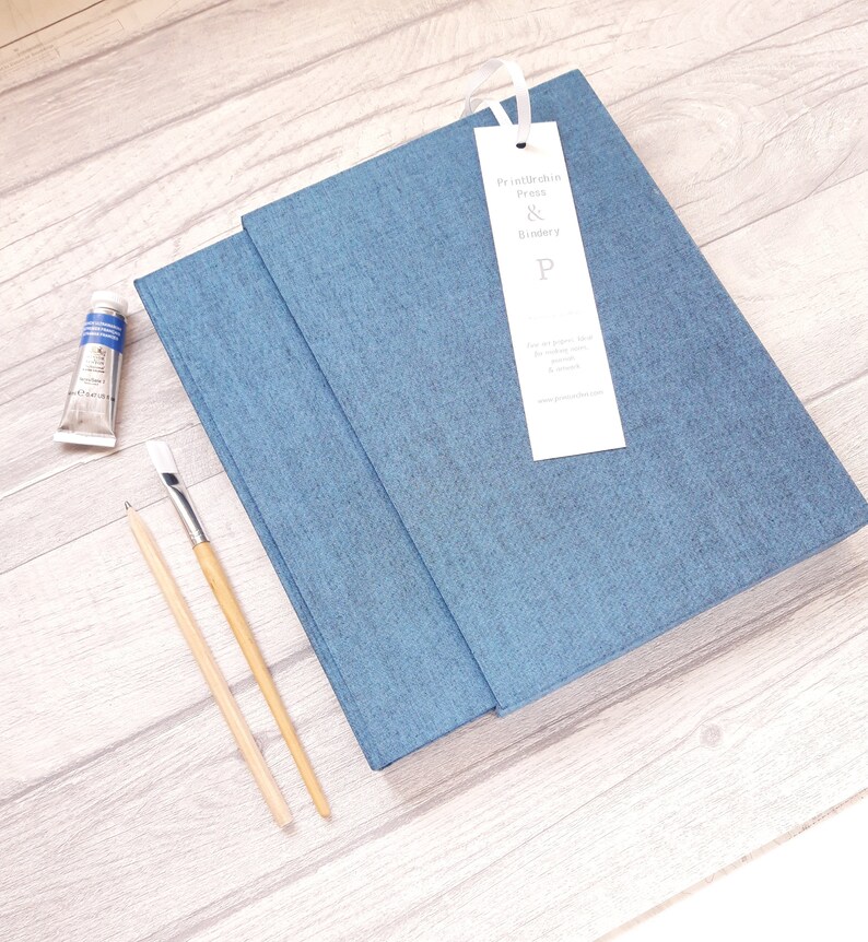 May include: A blue fabric-covered journal with a white ribbon bookmark featuring the text Printurchin Press & Bindery P. Beside the journal are a small tube of blue paint, a wooden paintbrush, and a wooden pencil, all arranged on a light wood surface.