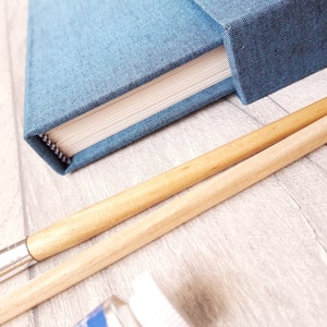 May include: A blue fabric-covered notebook with white pages and a square spine, positioned next to two wooden-handled paintbrushes with silver ferrules and a small portion of a white paint bottle cap visible at the bottom of the frame.