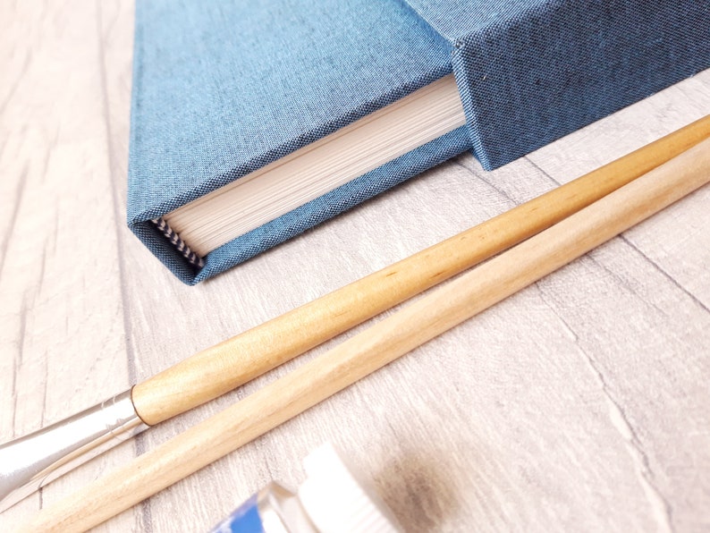 May include: A blue fabric-covered notebook with white pages and a square spine, positioned next to two wooden-handled paintbrushes with silver ferrules and a small portion of a white paint bottle cap visible at the bottom of the frame.