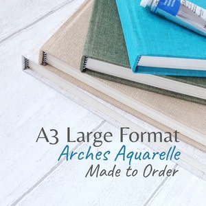 Pode incluir: Uma pilha de blocos de papel aquarela A3 em bege, verde e azul-turquesa. Um tubo de tinta French Ultramarine está em cima. O texto na imagem diz "A3 Large Format Arches Aquarelle Made to Order."