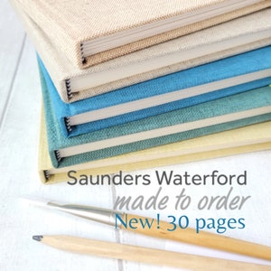 May include: A stack of hardcover journals in various colors, including beige, blue, and teal. The text "Saunders Waterford made to order New! 30 pages" is visible. A paintbrush and pencil are in the foreground.