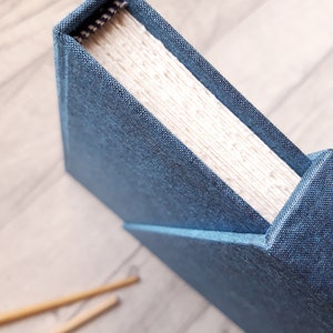 May include: A hardbound book featuring a textured dark blue fabric cover. The spine shows a visible woven head-and-tail band with a black and white pattern. The pages are cream-colored and appear thick, with a visible grain texture along the top edge.