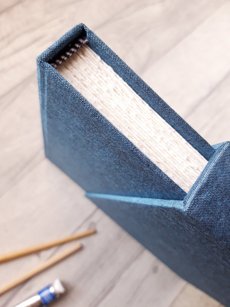 May include: A hardbound book featuring a textured dark blue fabric cover. The spine shows a visible woven head-and-tail band with a black and white pattern. The pages are cream-colored and appear thick, with a visible grain texture along the top edge.