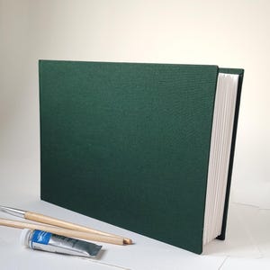 May include: A dark green sketchbook with a hard cover and white paper pages. Two paintbrushes and a tube of blue paint are in front of the sketchbook, suggesting art supplies. The sketchbook is upright, with the pages visible.