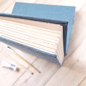 May include: A blue fabric-bound journal with thick, cream-colored pages featuring rough, deckled edges. The book is positioned at an angle, revealing the textured weave of the cover material and the layered, uneven texture of the paper sheets.