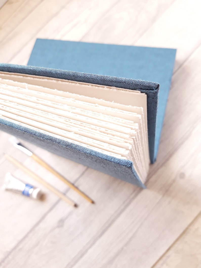 May include: A blue fabric-bound journal with thick, cream-colored pages featuring rough, deckled edges. The book is positioned at an angle, revealing the textured weave of the cover material and the layered, uneven texture of the paper sheets.