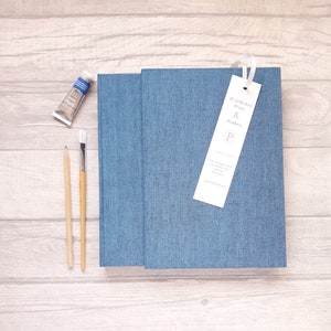 May include: A blue fabric-covered book with a white bookmark featuring the text Printurchin Press & Bindery P. Beside the book are a small tube of blue paint, a wooden pencil, and a paintbrush with a white bristle tip and a light wooden handle.