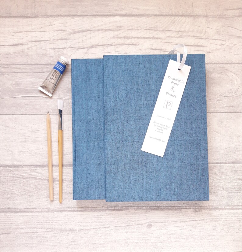 May include: A blue fabric-covered book with a white bookmark featuring the text Printurchin Press & Bindery P. Beside the book are a small tube of blue paint, a wooden pencil, and a paintbrush with a white bristle tip and a light wooden handle.