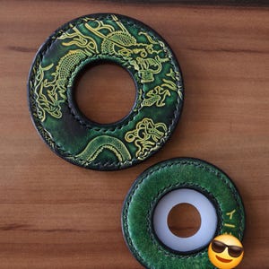 Puede incluir: Dos objetos circulares de cuero en tonos verdes y negros. La pieza más grande presenta un diseño de dragón en relieve en amarillo y verde. La pieza más pequeña tiene una superficie verde lisa con un inserto central blanco y un pequeño carácter japonés amarillo.