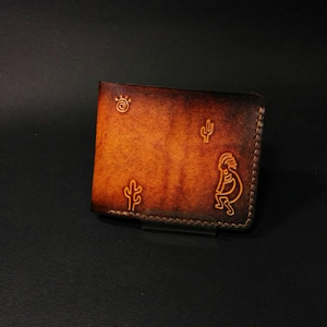 May include: A brown leather wallet with a desert theme. The wallet features a sun, cactus, and Kokopelli design. The edges are stitched with a contrasting thread. The wallet is a bi-fold style.
