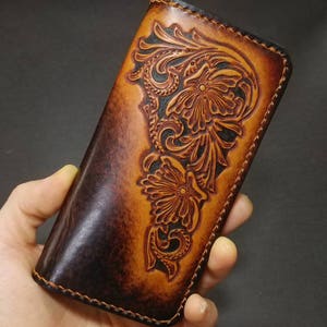 Puede incluir: Una cartera de cuero hecha a mano con un diseño floral en relieve. La cartera presenta un degradado de tonos marrones, desde el marrón oscuro hasta un bronceado más claro, con intrincados patrones florales. Los bordes están cosidos con un hilo en contraste.