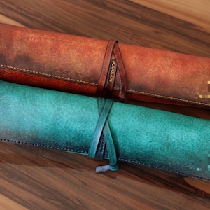 May include: Two leather pen rolls, one in a reddish-brown hue and the other in teal. Both are rolled up and secured with leather straps. The pen rolls have a distressed finish and are likely handmade. The rolls are on a wooden surface.