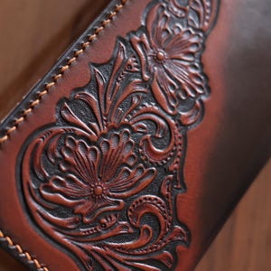 May include: A brown leather wallet featuring a deep-relief floral carving with intricate scrollwork and petals. The leather has a two-tone finish with dark brown recessed areas and lighter brown raised surfaces, finished with visible tan stitching.