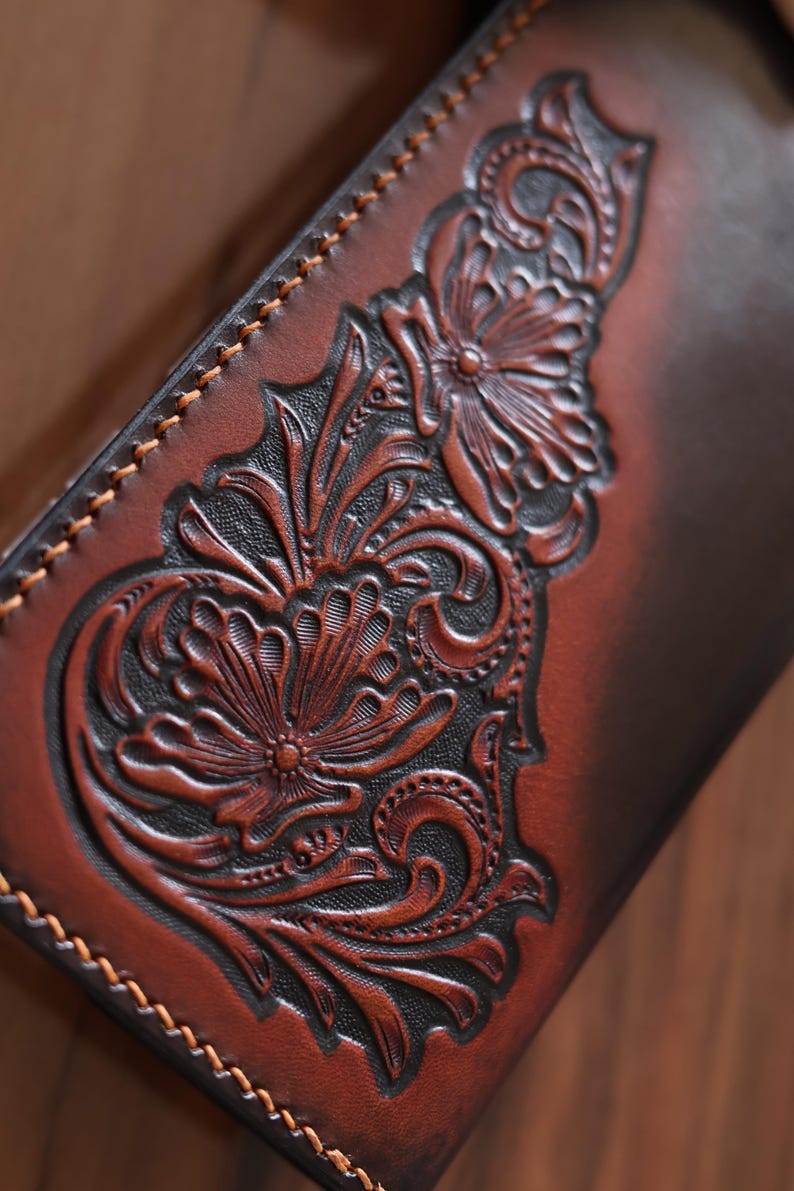 May include: A brown leather wallet featuring a deep-relief floral carving with intricate scrollwork and petals. The leather has a two-tone finish with dark brown recessed areas and lighter brown raised surfaces, finished with visible tan stitching.