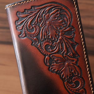May include: A rectangular brown leather wallet featuring a dark-toned, hand-tooled floral pattern with intricate carved details. The edges are finished with thick, light-colored stitching that contrasts against the deep, gradient-dyed leather surface.