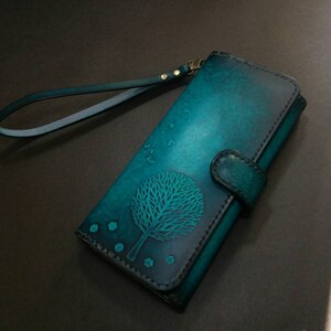 May include: Teal and navy blue leather wallet with a tree design. The wallet has a wrist strap and a snap closure. The tree design is embossed on the front of the wallet. The wallet is rectangular and has a smooth finish.