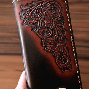 May include: A rectangular leather wallet featuring a dark brown and tan gradient finish. The surface displays an intricate, hand-tooled floral pattern with deep carvings. The edges are secured with thick, light-colored stitching along the perimeter.