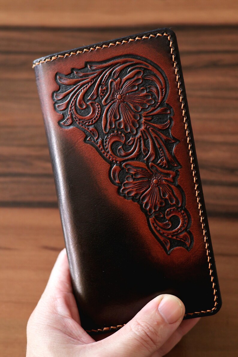 May include: A rectangular leather wallet featuring a dark brown and tan gradient finish. The surface displays an intricate, hand-tooled floral pattern with deep carvings. The edges are secured with thick, light-colored stitching along the perimeter.