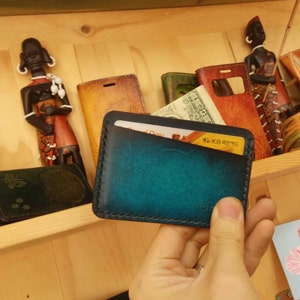 May include: A teal and black leather cardholder with a gradient effect, holding a credit card and cash. The wallet is displayed on a wooden shelf with other leather goods and decorative figurines.