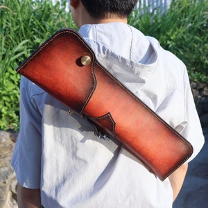 May include: A brown leather quiver with a shoulder strap. The quiver has a rich, reddish-brown color with darker edges and a brass button closure. It is designed to hold arrows and is carried over the shoulder.