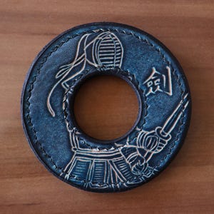 Peut inclure: Une tsuba circulaire en cuir bleu foncé ornée d'un pratiquant de kendo en relief portant un masque de protection men et tenant un sabre shinai. Le motif inclut le kanji japonais pour sabre et des coutures décoratives sur les bords.