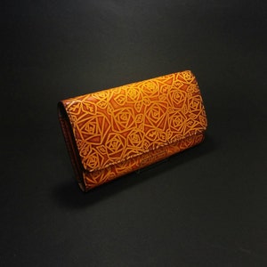 May include: A brown leather wallet with a rose pattern embossed on the exterior. The wallet is rectangular with a fold-over flap. The roses are a lighter shade of brown than the background, creating a contrast. The wallet is on a black surface.