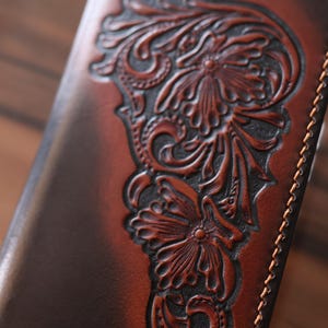 May include: A brown leather wallet featuring a deep, embossed floral pattern with intricate scrollwork and petals. The leather has a gradient finish, transitioning from dark brown edges to a lighter center, complemented by light tan stitching along the side.