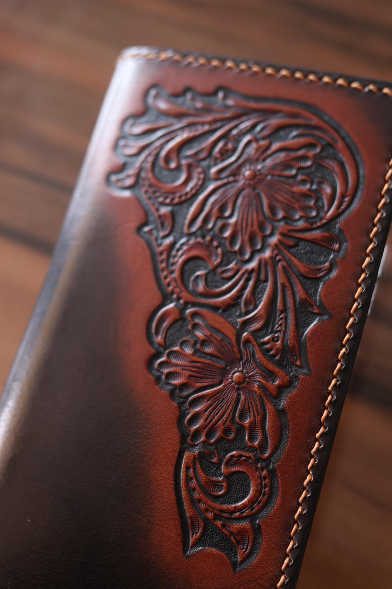 May include: A brown leather wallet featuring a deep, embossed floral pattern with intricate scrollwork and petals. The leather has a gradient finish, transitioning from dark brown edges to a lighter center, complemented by light tan stitching along the side.