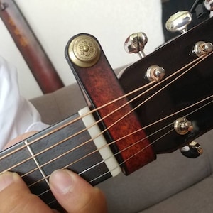 May include: A close-up of a guitar neck with a capo attached. The capo is made of brown leather with a decorative gold-colored metal button. The guitar strings are visible, and the fretboard is dark brown.