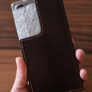 May include: A dark brown leather phone case featuring a rectangular cutout for the camera module. The case is constructed with visible orange stitching along the edges and includes a clear plastic protective cover over the camera lens area.