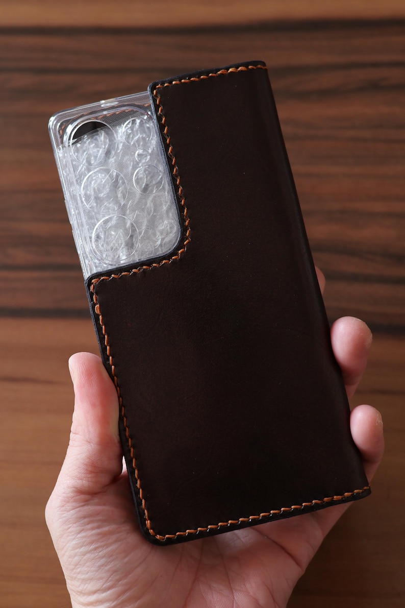 May include: A dark brown leather phone case featuring a rectangular cutout for the camera module. The case is constructed with visible orange stitching along the edges and includes a clear plastic protective cover over the camera lens area.