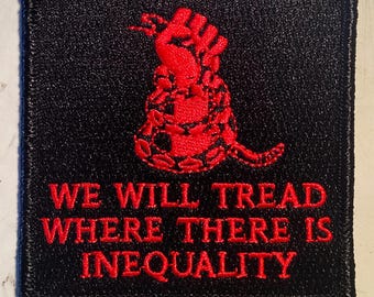 Puede incluir: Un parche negro con un diseño de serpiente y puño rojos. El texto "WE WILL TREAD WHERE THERE IS INEQUALITY" está bordado en rojo.