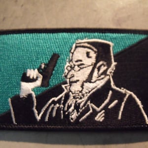 May include: Black and white embroidered patch of a person holding a gun, with a teal green background and a black and white diagonal stripe.