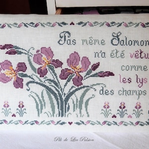 Puede incluir: Una almohada de lino blanco con un diseño de punto de cruz con iris púrpuras y una cita en francés: "Pas même Salomon n'a été vêtu comme les lys des champs." La cita está bordada en hilo rosa y verde. La almohada está ribeteada con un borde verde y rosa.