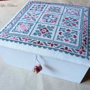 Peut inclure: Une boîte en bois blanche avec un couvercle à charnière. Le couvercle est recouvert d'un tissu blanc avec un motif de point de croix présentant une variété de motifs floraux roses et bleus. Le texte "Pili de Lin Pulsion" est brodé dans le coin supérieur gauche du couvercle. Une petite bobine de fil en bois est attachée à la boîte avec une ficelle blanche.
