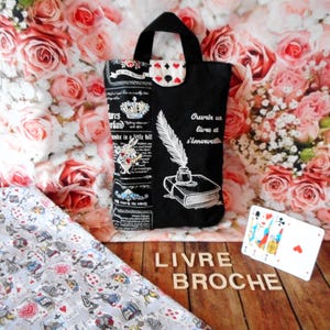 Peut inclure: Sac fourre-tout noir avec un motif blanc d'Alice au pays des merveilles et le texte "Ouvrir un livre et s'émerveiller". Le sac a une poignée noire et une petite poche avec un motif de cœur. Un jeu de cartes et les mots "LIVRE BROCHE" sont également visibles.