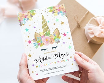 May include: A white invitation card featuring a unicorn design, floral decorations, and gold stars. The text reads "Anna Myer Magical 1st Birthday." The card is held by hands, with a gift box and ribbon in the background.