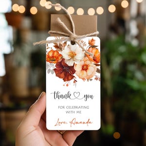 Może przedstawiać: Biała etykieta prezentowa z kwiatowym wzorem w odcieniach pomarańczy, czerwieni i bieli. Etykieta zawiera napis "thank you" i "for celebrating with me" z imieniem "Amanda" poniżej. Etykieta jest związana sznurkiem.