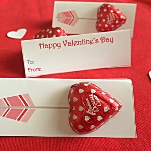 May include: Two white Valentine's Day cards with a red heart-shaped chocolate candy on each. The cards have a pink and gray arrow design and the text "Happy Valentine's Day", "To:", and "From:".