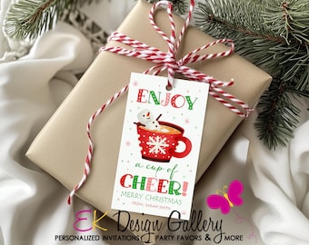 May include: A wrapped gift with a red and white twine bow and a Christmas tag. The tag reads "ENJOY a cup of CHEER! MERRY CHRISTMAS" and features a snowman in a red mug. The gift is on a white surface with evergreen branches.