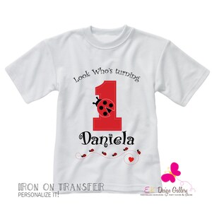 May include: White t-shirt with a red number one and a ladybug design. The text "Look Who's turning" and "Daniela" are printed on the shirt.